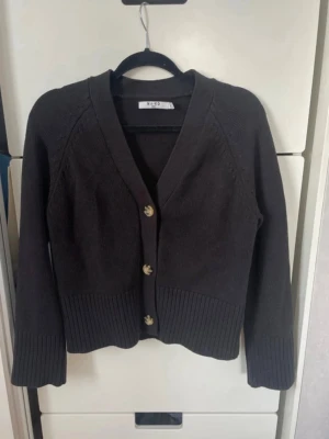 Svart kofta från NA-KD med knappar - Snygg svart kofta från NA-KD med tre dekorativa knappar framtill. Koftan har v-ringning, långa ärmar och bred ribbstickning vid ärmslut och nederkant. Perfekt att slänga över en topp för en stilren look.