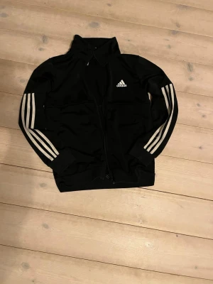Svart Adidas träningsjacka - Svart träningsjacka från Adidas med klassiska vita ränder längs ärmarna och vit Adidas-logga på bröstet. Jackan har dragkedja framtill, hög krage och ribbade muddar. Perfekt för sport eller chill dagar.