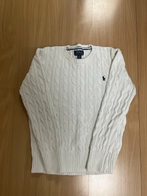 Vit kabelstickad tröja Polo Ralph Lauren - Kabelstickad vit tröja från Polo Ralph Lauren med rund halsringning och klassisk marinblå logga broderad på bröstet. Tröjan har ribbade muddar vid ärmslut och nederkant och är tillverkad i mjukt material. Perfekt för en clean och stilren look.