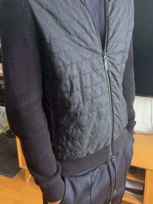 Svart J Lindeberg cardiganjacka - Snygg svart cardiganjacka och stickade ärmar. Jackan har dragkedja framtill och ribbade muddar vid ärmslut och midja. Perfekt för lager-på-lager och en stilren look. Storlek S men lite stor för att vara S