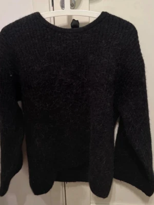 Svart stickad tröja med rosett - Mysig svart stickad tröja med lång ärm och rund halsringning. Baksidan har en djupare urringning och en dekorativ svart rosett i nacken. Tröjan har en fluffig struktur och ribbstickade muddar vid ärmslut och nederkant.