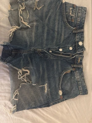 Blå slitna jeansshorts med knappar - Säljer ett par blå jeansshorts från ZARA med hög midja och slitna detaljer. Shortsen har flera knappar framtill och klassiska fickor både fram och bak. Perfekta för en avslappnad och trendig sommarlook ☺️dom är användna men fint skick 