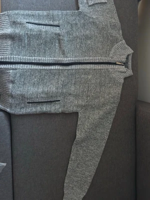 Grå stickad ziptröja med fickor - Snygg gråmelerad stickad tröja med dragkedja framtill och två fickor med svarta detaljer. Tröjan har ribbstickade muddar vid ärmslut och nederkant samt hög krage. Perfekt för lager på lager och enkel att matcha.