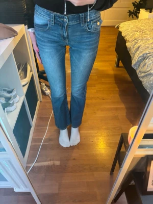 Blå raka jeans med fickdetalj - Säljer ett par klassiska blå jeans med rak passform och normal midja. Jeansen har snygga fickor bak med knappdetalj och är i ett mjukt denimtyg. Perfekta till vardagslooken och lätta att matcha med olika stilar.