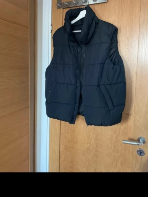 Svart dunväst från 157 - Snygg svart dunväst från 157 med hög krage och dragkedja framtill. Västen har en puffig look och är perfekt för lager-på-lager-stil. 