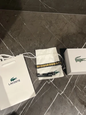 Svart och guld armband från Lacoste - Snyggt armband från Lacoste i svart och guld metall med länkdesign och små krokodilmotiv på varje länk. Kommer med originalpåse, verktyg för justering och presentask. Perfekt accessoar för dig som gillar stilrena detaljer och exklusiv känsla.