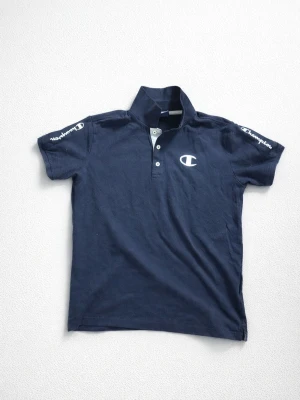 Mörkblå piké från Champion - Snygg mörkblå pikétröja från Champion med vit logga på bröstet och tryck på båda ärmarna. Klassisk krage och knappar framtill. Perfekt för en sportig och avslappnad stil.