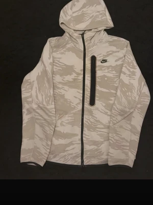 Nike camo hoodie med dragkedja - Snygg hoodie från Nike i beige och vitt camomönster. Tröjan har hel dragkedja, huva och en vertikal bröstficka med svart dragkedja. Nike-logga på bröstet. Perfekt för en sportig och avslappnad stil.