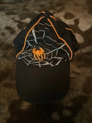 Svart keps med spindelmotiv - Säljer en svart keps med coolt spindelnätsmönster och en broderad orange spindel framtill. Kepsen har orangea detaljer längs sömmarna och böjd skärm. Perfekt för dig som gillar unika accessoarer med lite edge.
