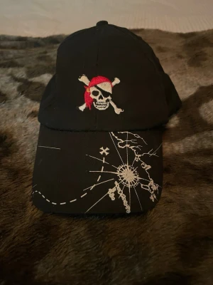 Svart keps med piratmotiv - Cool svart keps med broderad dödskalle och korslagda benknotor med röd bandana framtill. Skärmen har ett tryck av en skattkarta i vitt. Perfekt accessoar för dig som gillar äventyr och piratstil.