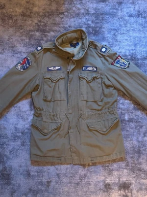 Militärinspirerad jacka från Polo Ralph Lauren - Militärgrön jacka från Polo Ralph Lauren med patchar på ärmar och bröst. Jackan har fyra fickor framtill, dragsko i midjan och ståkrage. Materialet är bomull och jackan har detaljer som knappar och dragkedja. Märket R.Lauren syns tydligt på bröstet.