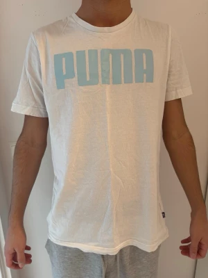 Vit PUMA t-shirt med blå logga - Snygg vit t-shirt från PUMA med stor ljusblå logga över bröstet. Klassisk passform och rund halsringning. Tillverkad i mjuk bomull som känns skön mot huden. Skulle säga den är lite mindre i storleken och passar en storlek S mer