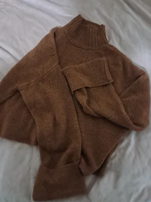 Brun stickad polotröja - Mysig brun stickad polotröja med hög krage och långa ärmar. Tröjan har en avslappnad passform och ribbstickade detaljer vid halsen och ärmsluten. Perfekt för kalla dagar och enkel att matcha med olika outfits.