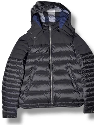 Svart burberry pufferjacka med huva - Snygg svart pufferjacka med avtagbar huva och dragkedja framtill. Jackan har quiltad design, två snedställda fickor och ett foder med rutigt mönster. Perfekt för kalla dagar och riktigt varm tack vare det tjocka materialet.