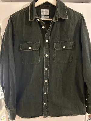 Mörk overshirt från Norse Projects - Snygg mörkgrå overshirt / jeansjacka från Norse Projects med klassisk krage, vita knappar och två bröstfickor med lock. Jackan har raka ärmar och kontrastsömmar som ger en clean look. Perfekt för dig som gillar stilrena och tidlösa plagg.Strokek S