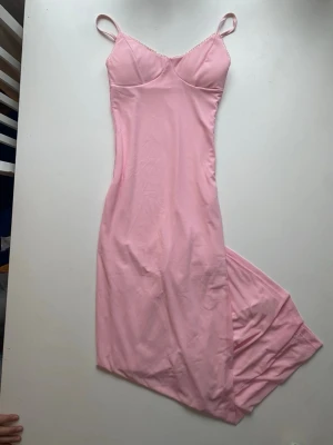 Ljusrosa långklänning med smala band - Säljer en ljusrosa långklänning med smala axelband och figurnära passform. Klänningen har en enkel, stilren design med lätt v-ringad byst och är tillverkad i ett mjukt, stretchigt material som ger en bekväm känsla.