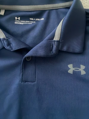 Marinblå piké från Under Armour - Snygg marinblå pikétröja från Under Armour med klassisk krage och knappslå. Tröjan har diskreta grå detaljer vid kragen och logga på bröstet. Tillverkad i ett lätt och funktionellt polyestermaterial som andas bra.