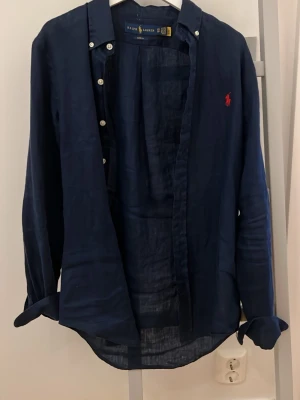 Mörkblå linneskjorta från Ralph Lauren - Klassisk mörkblå skjorta från Ralph Lauren i linne med knappar framtill och lång ärm. Ikonisk röd logga broderad på bröstet. Perfekt för en avslappnad men ändå stilren look. Perfekt nu till våren/sommaren. Använt den 2 gånger då den är för liten! Xs slimfit