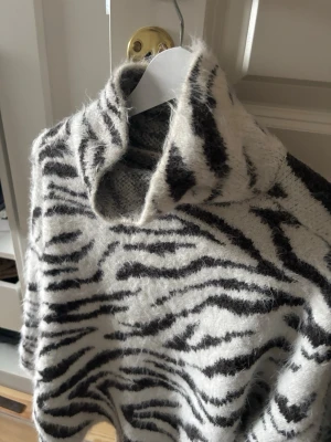Zebramönstrad stickad polotröja - Säljer en fluffig stickad polotröja med zebramönster i svart och vitt. Tröjan har hög krage och är gjord i ett mjukt, lurvigt material. 