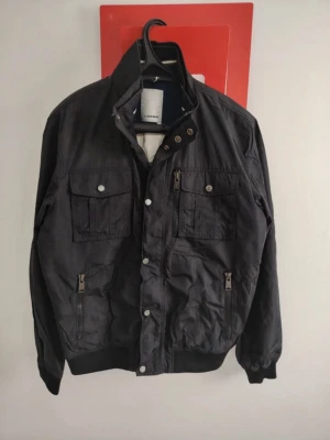 J.lindberg cardigan  - Snygg svart vindjacka med hög krage och flera praktiska fickor med dragkedja och knappar. Jackan har muddar vid ärmslut och nederkant samt knappslå framtill. Perfekt för dig som vill ha en stilren och funktionell jacka.