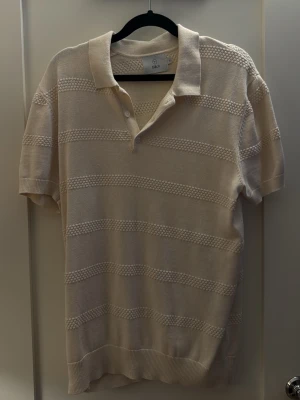 Beige stickad polotopp från Bläck - Säljer en beige stickad polotopp från Bläck med korta ärmar och diskret randigt mönster. Toppen har klassisk krage och knappar framtill för en clean look. Perfekt till jeans eller kjol för en chill och stilren vibe.