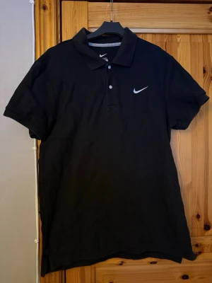 Svart piké t-shirt från Nike - Klassisk svart piké t-shirt från Nike med vit broderad logga på bröstet. Tröjan har korta ärmar, krage och knappar framtill. Perfekt för en sportig och stilren look.