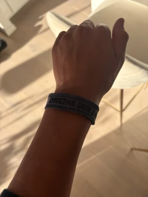 Grå Christian Dior armband med svart text - Snyggt mörkblått armband från Christian Dior med broderad logga och justerbar knytning med tofsar. Armbandet är gjort i textil och har en cool, avslappnad vibe som passar perfekt till din outfit.
