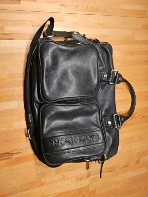 Svart datorväska från Björn Borg - Snygg svart datorväska från Björn Borg i läderimitation med två stora ytterfack framtill och justerbar axelrem. Väskan har rejäla handtag, dragkedjor i metall och vinrött innerfoder med flera innerfack för smart organisering. Perfekt för laptop och tillbehör.