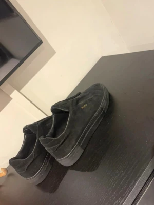 Svarta sneakers från Axel Arigato - Stilrena svarta sneakers från Axel Arigato i mocka med diskret guldfärgad logga på sidan. Lågt skaft och elastiska band över vristen för en clean look utan snörning. Perfekta för dig som gillar minimalistisk och modern stil.