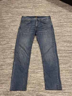 Blå raka jeans från Lee - Säljer ett par klassiska blå jeans från Lee med rak passform och fem fickor. Jeansen har en snygg tvättad look och Lee-lapp bak i midjan. Tillverkade i slitstarkt denimtyg, perfekta för en avslappnad stil.