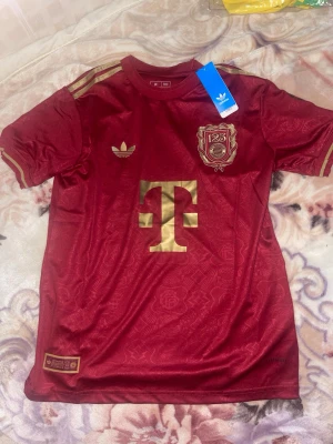 Bayern München tröja Adidas - Säljer en bordeauxröd Bayern München jubileumströja från Adidas med gulddetaljer och klubbmärke. Tröjan har korta ärmar, rund hals och är i slim fit-modell. Materialet är lätt och andas, perfekt för träning eller match. Priset kan diskuteras