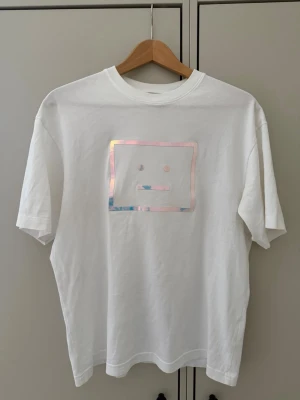 Acne Studios  - Vit t-shirt från Acne Studios med ett holografiskt ansikte-tryck på bröstet. Klassisk passform och rund halsringning. Tillverkad i 100% bomull för en mjuk och bekväm känsla. Perfekt för dig som gillar minimalistisk och modern stil.