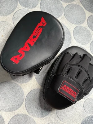Svarta fokusmittsar från Askari - Säljer ett par svarta fokusmittsar från Askar med röd logga. Tillverkade i slitstarkt konstläder och har vadderad insida för bra stötdämpning. Perfekta för boxningsträning eller kampsport. Snygg design och bekväm passform.