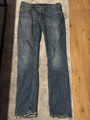 Blå nudie jeans med slitningar - Säljer ett par klassiska blå jeans med raka ben och lätt slitna detaljer. Jeansen har fem fickor, normal midja och kontrastsömmar. Perfekta för en avslappnad och trendig look. Materialet är denim i bomull.