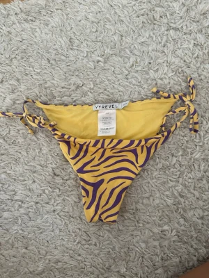 Gul-lila zebramönstrad bikini från Ivyrevel - Säljer en snygg bikiniunderdel från Ivyrevel i storlek 34. Den är gul med lila zebramönster och har knytband i sidorna för perfekt passform. Materialet är mjukt och stretchigt, perfekt för strandhäng eller poolparty.