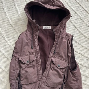 Brun väst med huva från Stone Island - Schysst brun väst från Stone Island med huva och dragkedja. Västen har två stora bröstfickor med knappar och detaljer i svart. Insidan är fodrad i mjuk fleece och det klassiska Stone Island-märket sitter på sidan. Perfekt lager på lager-plagg. Söker byte mot annat Stone Island eller sälja 