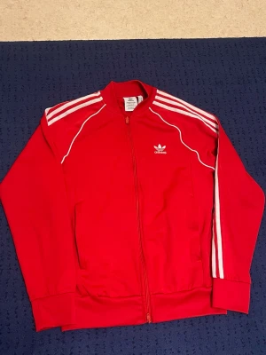 Röd Adidas tröja med dragkedja - Röd tröja från Adidas med dragkedja framtill och vita klassiska ränder på ärmarna. Tröjan har ribbade muddar, två fickor och Adidas logga på bröstet. Använt ett par gånger.