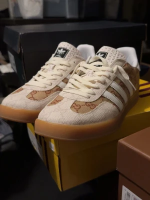 Adidas x Gucci beige gazelle  - Snygga sneakers i beige och brunt från Adidas x Gucci med klassiska vita ränder på sidan och gummisula. Ovandelen har Guccis GG-mönster i canvas och detaljer i vitt läder. Skorna har vita snören och Adidas-logga på plösen. Perfekt för dig som gillar exklusiva samarbeten.