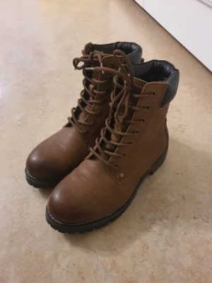 Bruna snörade kängor med svart detalj - Säljer ett par bruna kängor med snörning och grov sula. Kängorna har en svart vadderad kant upptill och är tillverkade i ett läderliknande material. Perfekta för dig som gillar en klassisk och robust stil.