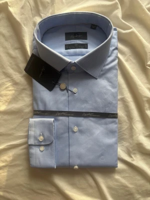 Ljusblå skjorta från Dressmann Signature - Klassisk ljusblå skjorta från Dressmann Signature med italienskt tyg. Skjortan har knappar framtill och på manschetten, samt en stilren krage. Perfekt för dig som vill ha en clean och tidlös look.