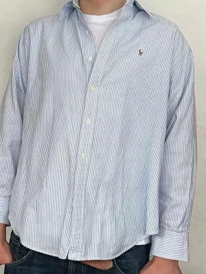 Blårandig skjorta från Polo Ralph Lauren - Klassisk blårandig skjorta från Polo Ralph Lauren med vit bas och tunna blå ränder. Skjortan har lång ärm, knappar framtill och den ikoniska broderade loggan på bröstet. Tillverkad i mjuk bomull för en bekväm känsla. Nypris 1700. Strl 12. Jag är 188 och väger 80 och den är bara lite kort i armarna 🍾💵