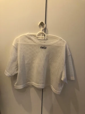 Vit crop top från AWCO - Säljer en vit nät croptop från AWCO. Tröjan har korta ärmar och är gjord i ett luftigt, genomskinligt nätmaterial som passar perfekt till träning eller som ett lager över sport-bh. Enkel och sportig design med svart logga 