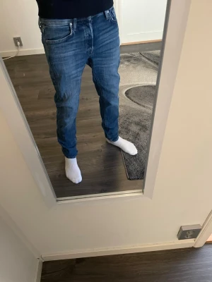 Blå jeans från Jack & Jones - Säljer ett par klassiska blå jeans från Jack & Jones med normal passform och snygga slitningar. Jeansen har fyra fickor, gul söm och är tillverkade i denim. Perfekta för en avslappnad och trendig stil. Pris kan diskuteras 