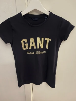 Svart GANT t-shirt med guldtext - Snygg svart t-shirt från GANT med glittrig guldtext 'GANT New Haven' på bröstet. Klassisk rund halsringning och korta ärmar. Tillverkad i mjuk bomull som känns skön mot huden. Perfekt för en stilren och trendig look.