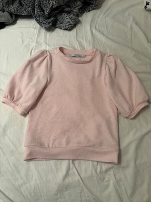 Ljusrosa topp med puffärm från H&M - Supersöt ljusrosa topp från H&M med puffiga korta ärmar och rund halsringning. Tillverkad i mjukt fleecematerial som känns skönt mot huden. Enkel men trendig design som passar perfekt till jeans eller kjol.