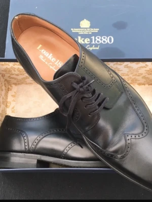 Svarta Loake 1880 läderloafers - Säljer ett par klassiska svarta loafers från Loake 1880 i äkta läder. Skorna har snygga broguedetaljer, rundad tå och eleganta snörningar. Perfekta för dig som vill ha en stilren och tidlös look. Tillverkade i England.