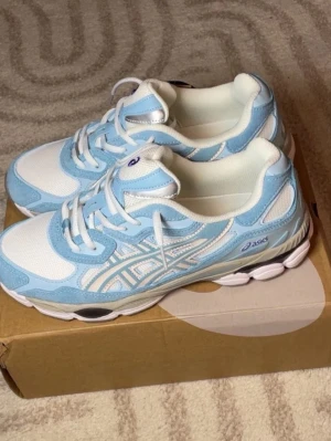 Asics vita och ljusblå sneakers - Snygga sneakers från Asics i vitt och ljusblått med klassiska ränder på sidan. Ovandelen är i mesh och syntetmaterial för bra andningsförmåga och komfort. Sulan är vit och grå med bra grepp. Perfekta för dig som gillar sportig och fräsch stil.