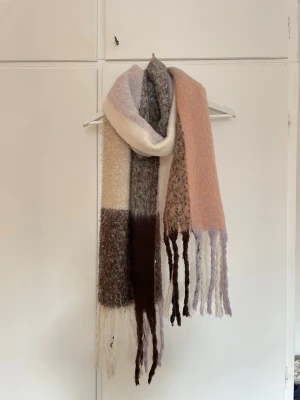 Flerfärgad lång halsduk med fransar - Mysig halsduk med nyanser av beige, brun, lila och rosa. Lång modell med grovstickad känsla och långa fransar i ändarna. Perfekt accessoar för att få lite extra färg till din outfit.