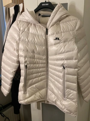 Vit dunjacka från J.Lindeberg - Snygg vit dunjacka från J.Lindeberg med huva och två dragkedjeförsedda fickor framtill. Jackan har ett diskret logotryck på bröstet och quiltad design med horisontella sömmar. Perfekt för kalla vinterdagar och riktigt lätt att bära. Helt ny med prislapp på. 