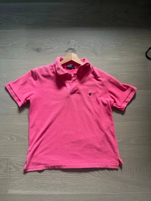 Rosa pikétröja från Polo Ralph Lauren - Klassisk rosa pikétröja från Polo Ralph Lauren med korta ärmar och krage. Tröjan har två knappar vid halsen och den ikoniska broderade loggan på bröstet. Tillverkad i mjuk bomull som känns skön mot huden.
